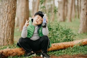 Young boy sitting outdoors holding a soft drink and making a peace sign in a forest.