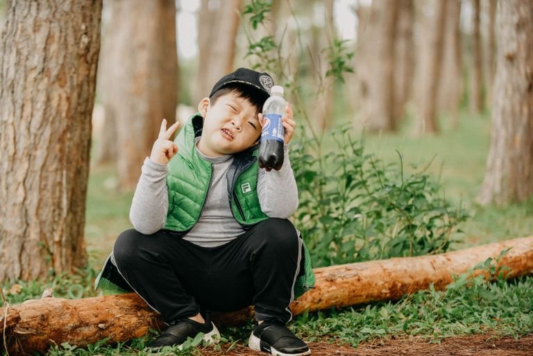 Young boy sitting outdoors holding a soft drink and making a peace sign in a forest.